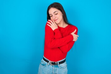 young brunette girl wearing red T-shirt against blue wall Hugging oneself happy and positive, smiling confident. Self love and self care