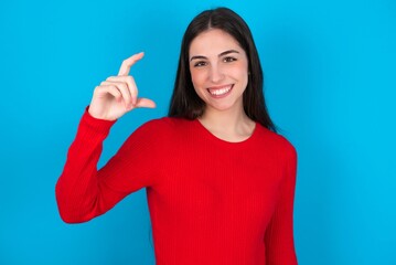 young brunette girl wearing red T-shirt against blue wall smiling and confident gesturing with hand doing small size sign with fingers looking and the camera. Measure concept