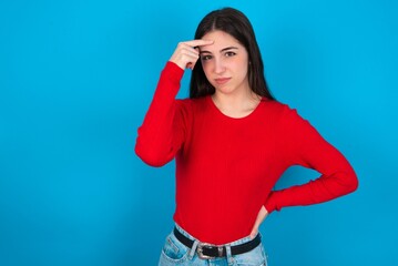 young brunette girl wearing red T-shirt against blue wall pointing unhappy at pimple on forehead, blackhead  infection. Skincare concept.