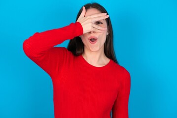 young brunette girl wearing red T-shirt against blue wall peeking in shock covering face and eyes with hand, looking through fingers with embarrassed expression.