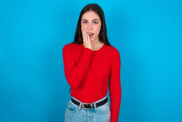 Shocked young brunette girl wearing red T-shirt against blue wall looks with great surprisment being very stunned, astonished with unexpected news, Facial expressions concept.