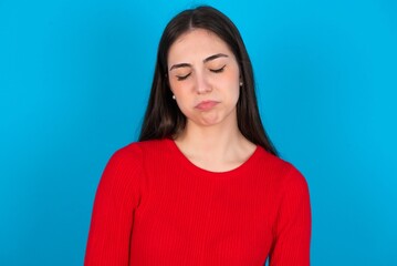 Obraz premium Dismal gloomy rejected young brunette girl wearing red T-shirt against blue wall has problems and difficulties, curves lower lip and closes eyes in despair, being in depression