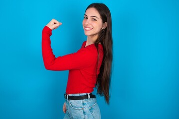 Portrait of powerful cheerful young brunette girl wearing red T-shirt against blue wall showing...