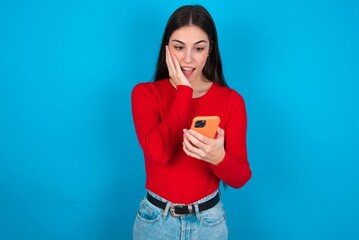 young brunette girl wearing red T-shirt against blue wall holding in hands cell reading sms using new app 5g