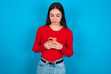 Excited young brunette girl wearing red T-shirt against blue wall winking and eye hold smart phone use read social network news