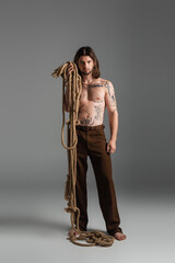 Barefoot and tattooed model holding rope on grey background.