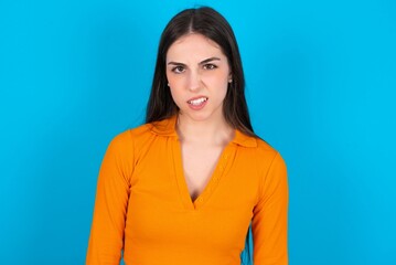 Portrait of dissatisfied young caucasian brunette girl wearing orange crop top against blue wall smirks face, purses lips and looks with annoyance at camera, discontent hearing something unpleasant