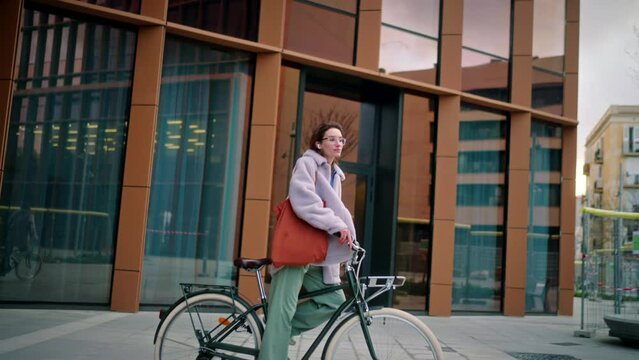 young trendy looking student wearing glasses cycling around the city near a modern urban business building. good vibe concept female hipster riding a green rental bike