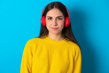 Serious displeased young brunette girl wearing yellow knitted sweater against blue wall looks puzzled at camera being angry wears stereo headphones listens music while walking at street
