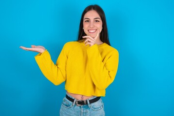 Positive young brunette girl wearing yellow knitted sweater against blue wall advert promo touch finger teeth