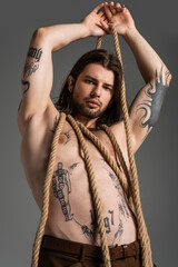 Low angle view of sexy tattooed man posing with rope isolated on grey.