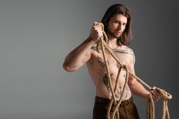Young long haired model with tattoo on body holding rope isolated on grey.