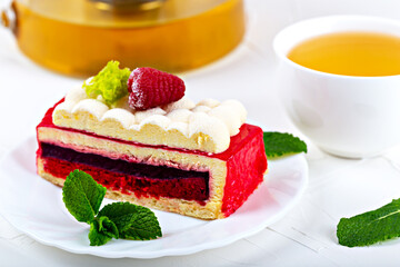 A piece of raspberry sponge cake with fluffy cream and mint on a white plate. Next to it is tea in a teapot and a cup