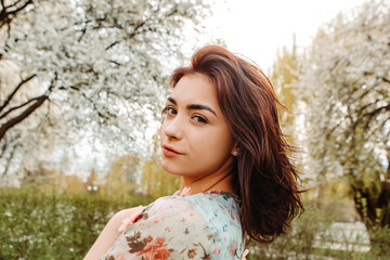 Naklejka premium Portrait of charming pretty woman dressed flowery dress posing near apple cherry tree blossoms blooming flowers in the garden park in early spring nature. Fashion, girl model with black hair