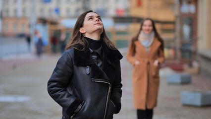 A friend comes up from behind her waiting girlfriend on a city street.