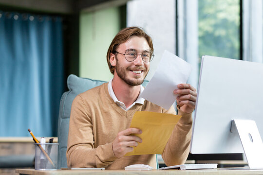 Young successful businessman in casual interior of office, blond man reading postal letter with good news, man happy with notification message in envelope, office worker with computer.