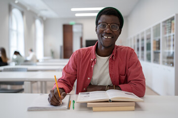 Happy carefree African American guy student age sits at desk in university auditorium. Cheerful young man is waiting for start of classes at city college for change profession or advanced training