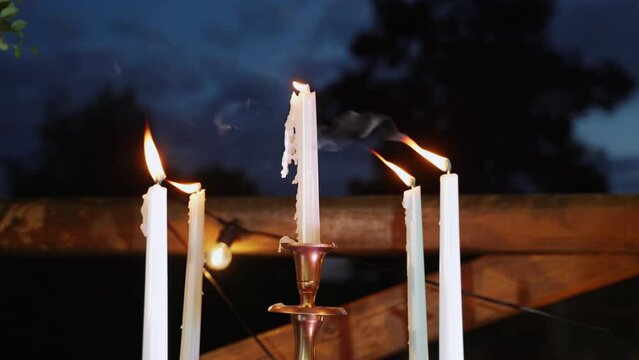Five mystical paraffin candles in candlestick near wooden fence with incandescent lamps burn at dusk outdoors. Wind stirs flame, melted wax flows down candles, silhouettes of trees in background