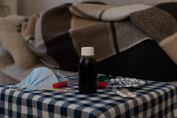 Disposable medical mask, thermometer, potions and pills on blurred background of person under the plaid on sofa