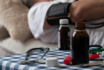 Phonendoscope, thermometer, potions and pills on blurred background of man on sofa at home
