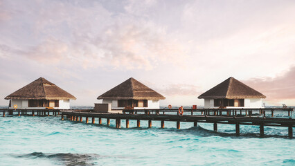 A wide-angle shot of a group of three overwater bungalows with thatched roofs connected via a wooden boardwalk in the background on a beautiful afternoon with some clouds in the lilac color sky