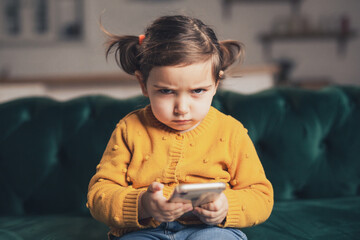 very angry little girl looking at the camera and holding device in her hands - obrazy, fototapety, plakaty