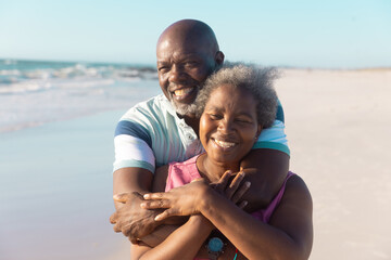Happy african american senior man embracing woman from back and enjoying at beach against sky
