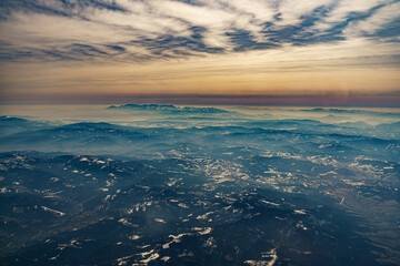 Fototapeta premium View from the airplane window at the setting sun and mountains crossed by clouds
