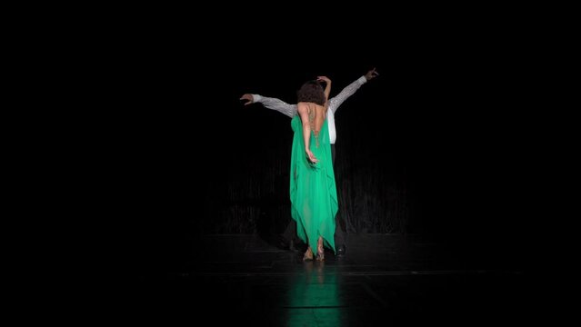 Dancer in green dress and her partner dance rumba in darkness. Wide hand movements accompany convergence and removal of performers, alternate rumba spiral turns. Girl performs three cucarachas and