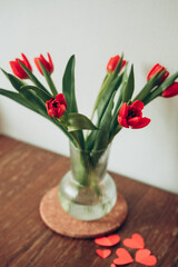 Red tulips flower bouquet in glass vase on table empty copy space.Fresh flowers on wooden background. red paper hearts