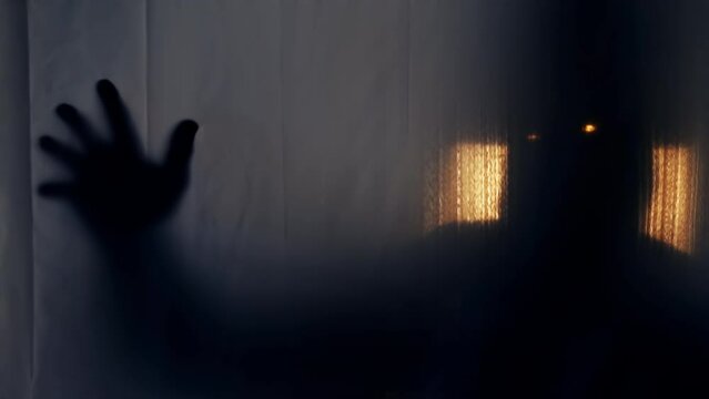 Silhouette of a man in a hood with glowing eyes behind a transparent curtain, pulling his hand into the camera. The concept of demons, paranormality, ghosts. 4k slowmo video with speed ramp effect.