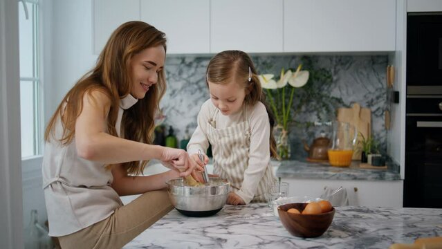 Cheerful family making dough at home. Girl helping millennial mom at kitchen