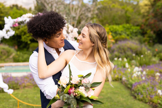 Happy diverse bride and groom holding bouquet embracing in garden at outdoor wedding