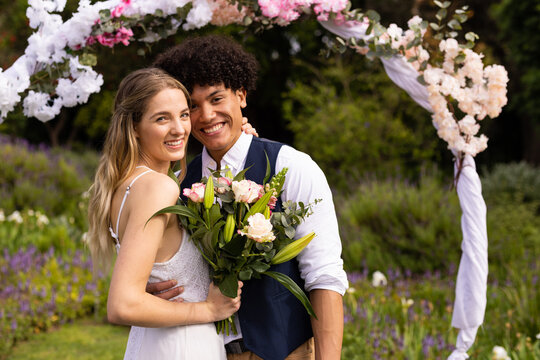 Happy diverse bride and groom holding bouquet embracing at outdoor wedding, copy space