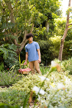 Vertical of biracial man walking in sunny garden with picnic basket, with copy space