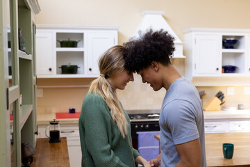 Happy diverse couple holding hands and touching heads, smiling in kitchen at home