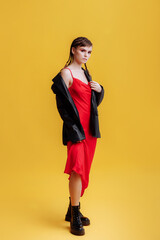 Beautiful female model in a red dress, black jacket and high-soled boots. Model portrait of a young beautiful fashion model posing on a yellow background. Bright makeup and braids.