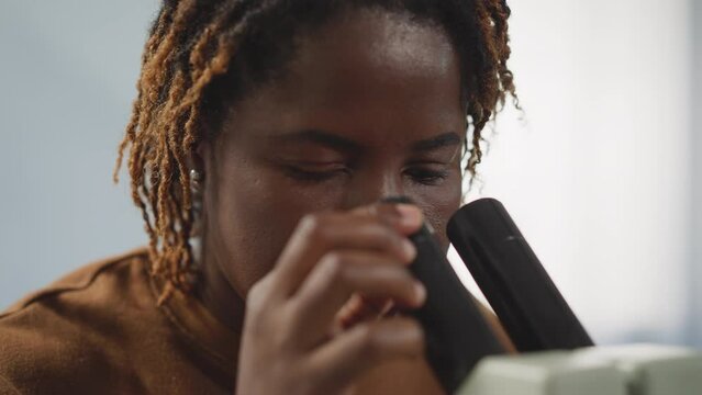 African American woman looks at viruses through microscope on blurred background. Female student provides medical research in modern laboratory closeup