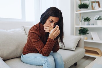 A woman looks at an engagement ring in her hand with sadness and tears, divorce and loss of a partner, breakup, unhappy heartbreak sitting at home on the couch