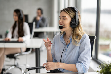 Young blonde haired woman in headset, sitting in office, working as operator in call center with her colleagues.