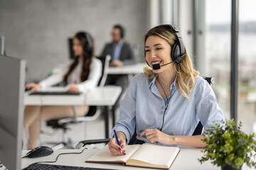 Young blonde woman working in call center, talking to client and taking notes to notebook.