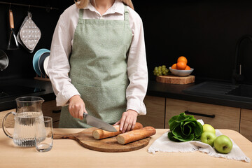 Woman in clean apron cutting baguette on wooden table at kitchen, closeup