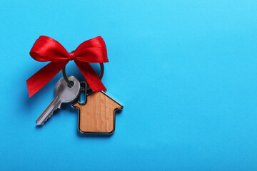 Key with trinket in shape of house and bow on light blue background, top view. Space for text. Housewarming party
