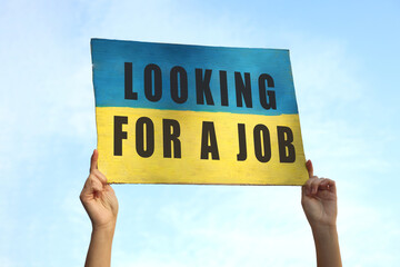 Unemployed due to war. Woman holding poster in colors of Ukrainian flag and phrase Looking For A Job against sky, closeup