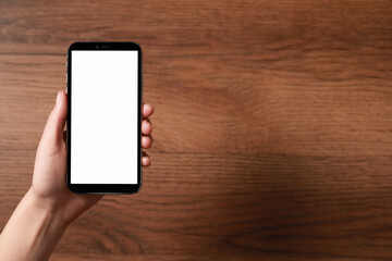 Woman holding smartphone with blank screen at wooden table, top view. Mockup for design
