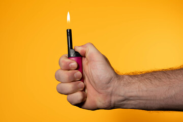 A man holding a lighter against a vibrant orange or yellow background. There is a flame coming out of the lighter.
