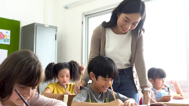 Asian school teacher assisting students in classroom. Young woman working in school helping boy with his writing, education, support, care.