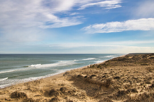 Under a partly cloudy sky on a snowless Winter day, ocean waves wash upon an empty beach beneath grassy sand dunes near Wellfleet, MA, on Cape Cod.