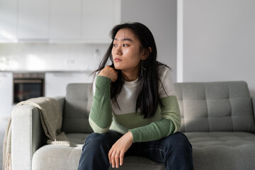Fototapeta premium Sad lonely young Asian woman having depression symptoms, sitting on sofa at home looking into distance, upset unhappy female worrying about money or family problems. Mental disorders, 