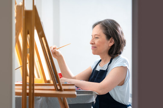 Happy female artist near workplace, smiling mature female artist in black apron standing with crossed arms near table with brushes and assorted paints while looking at camera in studio.